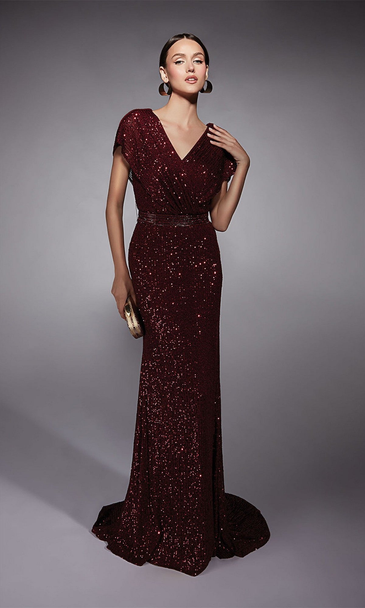 A woman in the Alyce 27708 long formal dress with short sleeves and a beaded waistband holds a gold clutch while posing against a plain gray background.